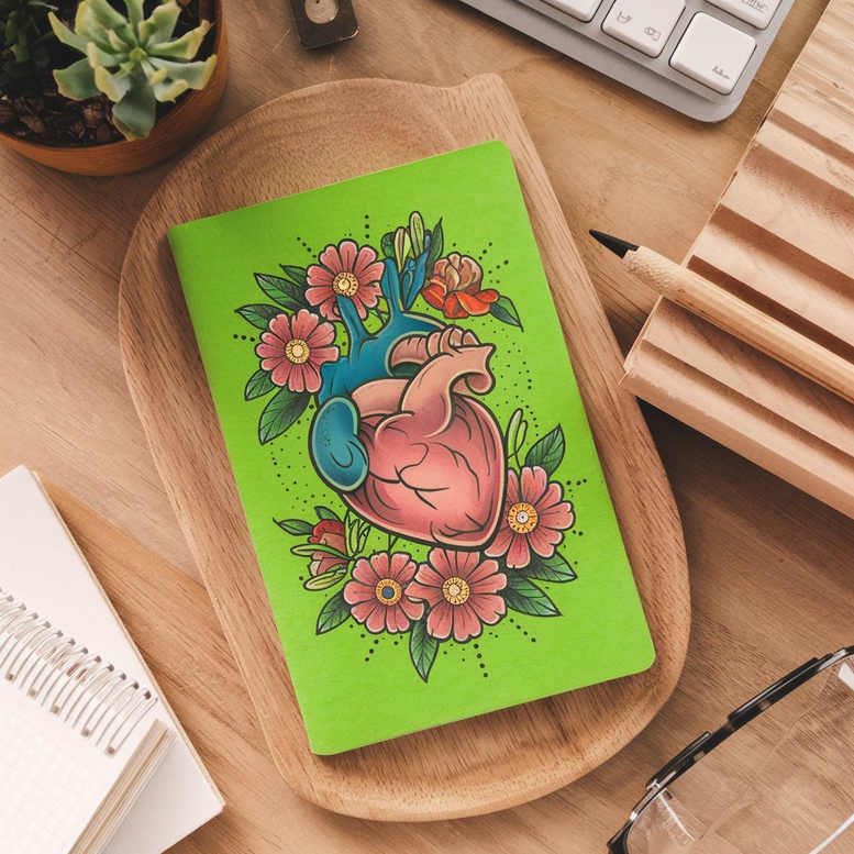 Notebook cover with blue-pink heart and pink flowers on vivid green; visible pencil, keyboard, succulent, and glasses; AI-generated tattoo project concept.