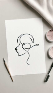 Paper with a black ink line drawing of a woman's profile; pencil, brush, and jar of paint visible; black and grey tattoo design concept, project suitable for cover-up.