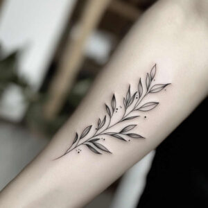 Black and grey fine-line botanical branch tattoo on forearm with leaves and small dots; project idea; not suitable for cover-up.